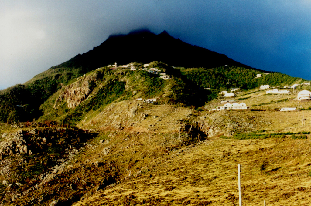 Mt. Scenery, the summit lava dome of Saba volcano, is seen from the airport on the NE side of the island.  The sun-dappled slopes in the foreground are the surface of a large andesitic lava flow that descends the NE flank of the volcano and forms the Flat Point Peninsula, on which the airport was constructed.  A steep switch-backed road extends from the airport to the village of Lower Hells Gate (center).  Saba is the northernmost active volcano of the West Indies. Photo by John Shepherd, 2000 (Seismic Research Unit, University of West Indies).