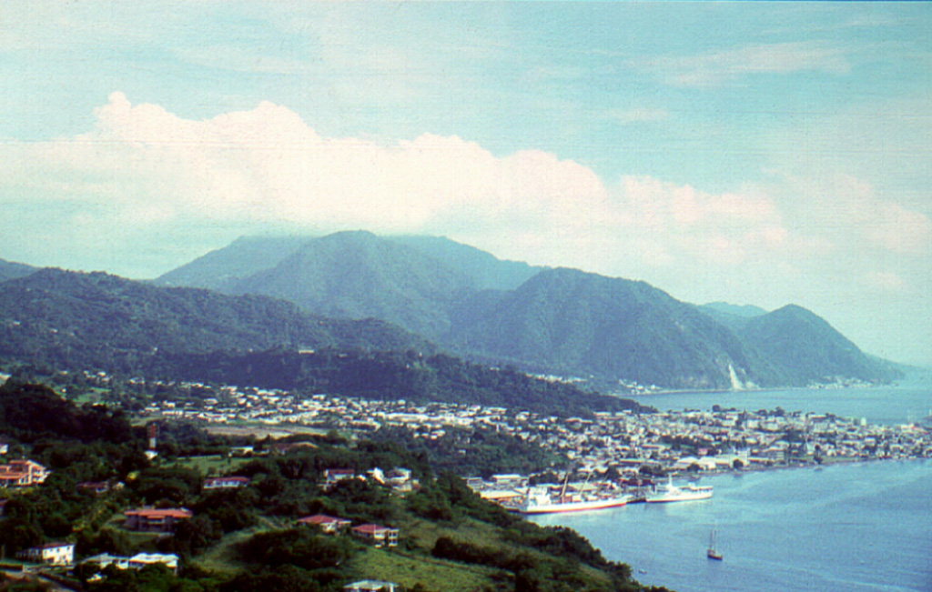 The cloud-capped summit of Morne Plat Pays stratovolcano lies on the horizon near the southern tip of the island of Dominica beyond the sprawling capital city of Roseau.  Three post-caldera lava domes (left-center to right) were formed north of a large arcuate depression open to the west that formed about 39,000 years ago.  The latest dated eruption at the Morne Plat Pays volcanic complex occurred from the Morne Patates lava dome about 1270 CE, although younger deposits have not yet been dated.   Photo by Paul Jackson, 1998 (Seismic Research Unit, University of West Indies).