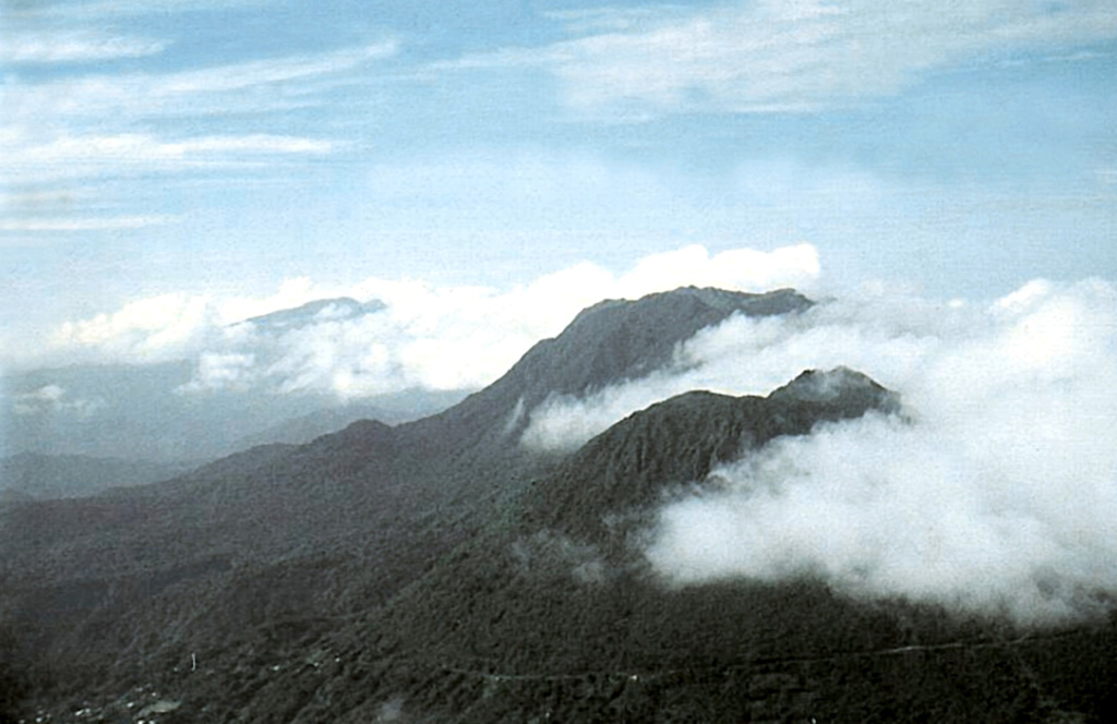 Two large lava domes, Micotrin (lower right) and Morne Trois Pitons (upper right), are located along the margin of a large semi-circular depression on the western coast of central Dominica NE of the capital city of Roseau.  The village of Laudat at the lower left lies along a road traversing the flanks of Micotrin on the way to the Atlantic coast.  The most recent dated dome-forming eruption at the Trois Pitons/Micotrin complex took place about 800 CE.  Morne Diablotins volcano lies in the clouds at the upper left.   Photo by Paul Jackson, 1998 (Seismic Research Unit, University of West Indies).