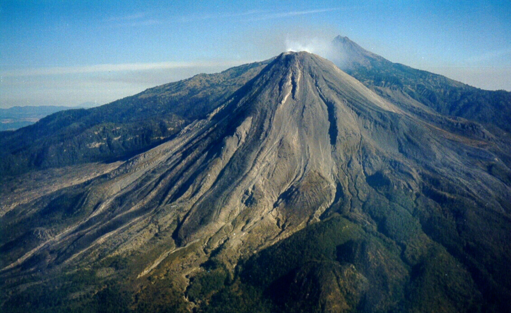 An aerial view of Colima's SW flank on 11 March 1999 shows recently erupted lava flows with lateral levees. Nevado de Colima appears to the right of the Volcán de Colima summit. Intermittent ash eruptions beginning in 1997 were followed by lava effusion, dome growth, pyroclastic flows, and more powerful explosive eruptions a year later. Photo by Juan Carlos Gavilanes, 1999 (Colima Volcano Observatory, University of Colima).