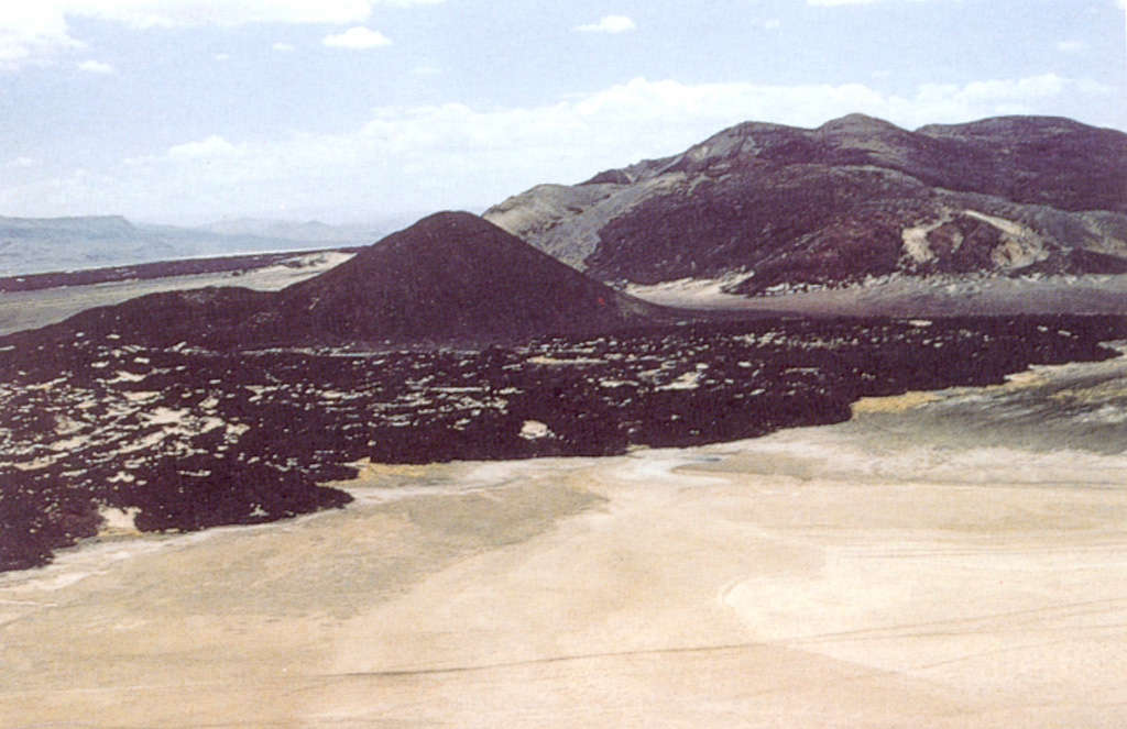 Basaltic lavas, scoria cones, and tuff deposits were erupted along the eastern base of Namarunu volcano along the axis of the East African Rift. This view looks S with the rift margin escarpment of the Tirr Tirr Plateau in the distance at the upper left. Numerous fissure-controlled scoria cones and lava flows, as well as partially or completely below lake level tuff cones, tuff rings, and pillow lavas were erupted along the flanks of the volcano. The youngest eruptions postdated the drying out of Lake Sugata about 3,000 years ago. Photo by Martin Smith, 1993 (copyright British Geological Survey, NERC).
