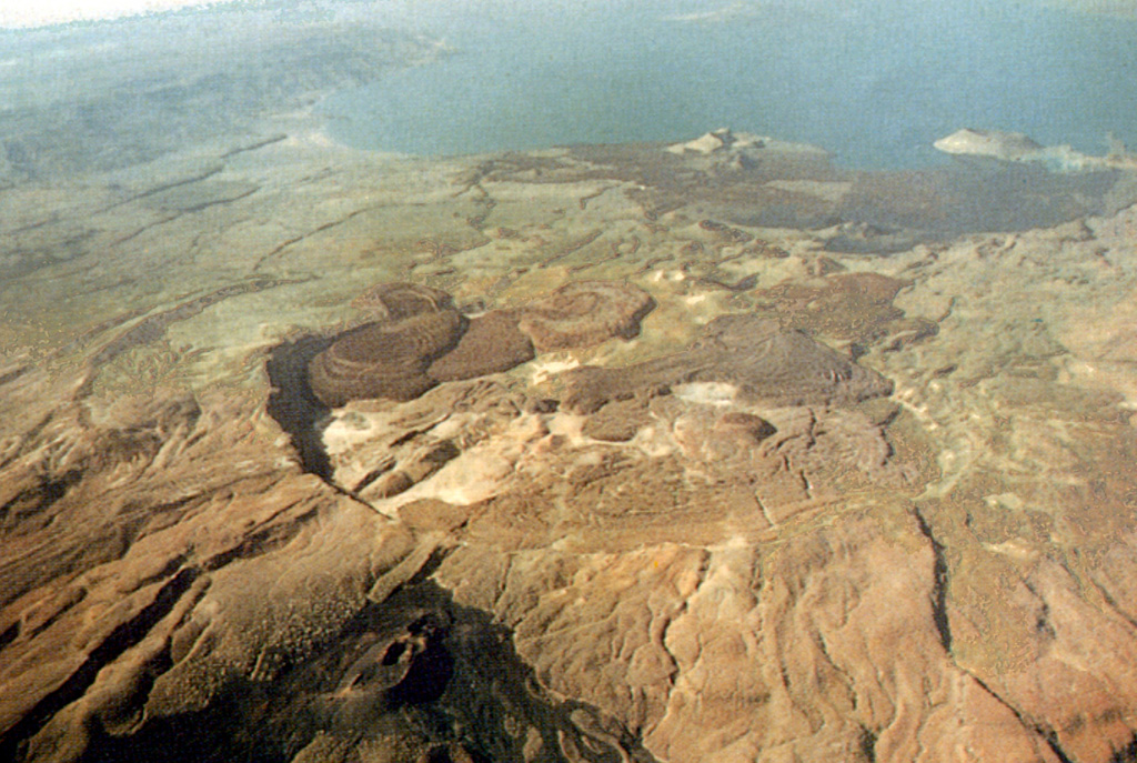 A northward-looking aerial view shows the summit caldera of Kakorinya volcano of The Barrier volcanic complex with Lake Turkana in the distance. The 3.8-km-wide summit caldera was formed about 92,000 years ago, and post-caldera lava domes and flows fill much of the caldera floor. Fresh-looking lava flows and tuff cones occur along the shores of Lake Turkana. Teleki's Cone on the northern flank and Andrew's Cone (lower left foreground) on the southern flank have been the source of historical lava flows. Photo by Martin Smith, 1993 (copyright British Geological Survey, NERC).