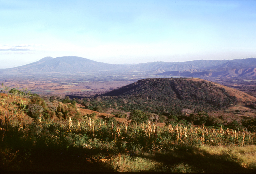 Cerro Alto (right) is a scoria cone on the SE flank of Coatepeque caldera, seen here from the southern caldera rim. Basaltic lava flows were erupted from the eastern side of the cone. Cerro Alto predates the formation of Coatepeque caldera and is overlain by deposits from the caldera-forming eruptions. San Salvador volcano is on the horizon to the east (left). Photo by Lee Siebert, 2002 (Smithsonian Institution).