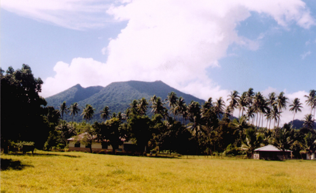 Gunung Ibu volcano on the NW coast of Halmahera Island rises beyond a small village on its WNW flank.  The summit of the stratovolcano is truncated by nested craters; the inner crater, 1 km wide and 400 m deep, contains several small crater lakes.  The outer crater is breached on the north side, forming the deep valley seen here on the left side of the summit ridge. Few eruptions are known in historical time from Ibu, although it was erupting at the time of this February 1999 photo. Photo by Agus Karim, 1999 (Volcanological Survey of Indonesia).