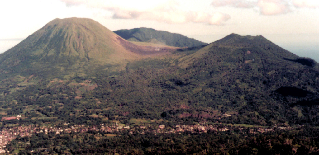 The twin volcanoes Lokon and Empung rise above the towns of Kakaskasen and Kinilow as viewed from the flanks of Mahawu volcano.  The more prominent Lokon volcano (left), is higher than Empung volcano (right) and lacks a summit crater.  Most historical eruptions from Lokon-Empung, one of the most active volcanoes on Sulawesi Island, have originated from Tompaluan crater, which can be seen surrounded by fresh ash deposits in the saddle between the two peaks.  Gunung Tetawiran rises beyond the saddle. Photo by Agus Solihin, 1998 (Volcanological Survey of Indonesia).
