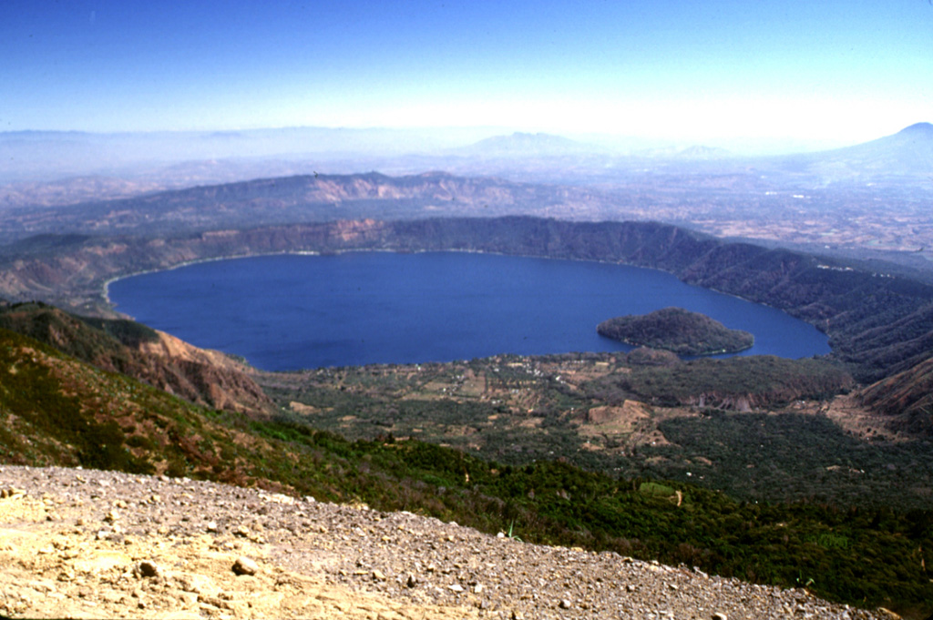 The 7 x 10 km Coatepeque caldera is seen here from the Santa Ana summit. The caldera formed during a series of major explosive eruptions between about 70,000 and 57,000 years ago. Post-caldera eruptions included the formation of basaltic scoria cones and lava flows near the western margin of the caldera and the extrusion of lava domes, including Cerro Grande, the island in the W side of the caldera lake. Photo by Lee Siebert, 2002 (Smithsonian Institution).
