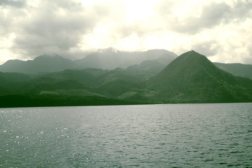 Cloud-draped Morne Diablotins volcano rises above the leeward coast of Dominica.  This little known volcano is the largest on Dominica and one of the largest of the Lesser Antilles.  Two coalescing lava domes form the summit of 1430-m-high Morne Diablotins, and precolumbian pyroclastic-flow aprons on the NW flank are relatively unmodified by erosion.  The sharp-topped peak in the right foreground is the Morne Espagnol (Barbers Rock) lava dome.  No historical eruptions are known from Morne Diablotins. Photo by Lee Siebert, 2002 (Smithsonian Institution).