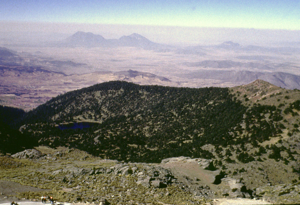 Glacial erosion has modified the summit of Cofre de Perote volcano. Summit lava flows show glacial striations, and a glacial tarn (left) is SW of the summit. Cofre de Perote overlooks the intermontaine Serdán-Oriental basin, which contains lava domes, tuff rings, lava flows, and scoria cones. The two Las Derrumbadas lava domes are in the distance and Cerro Pinto dome to their right are about 40 km SW. Photo by Lee Siebert, 1999 (Smithsonian Institution).