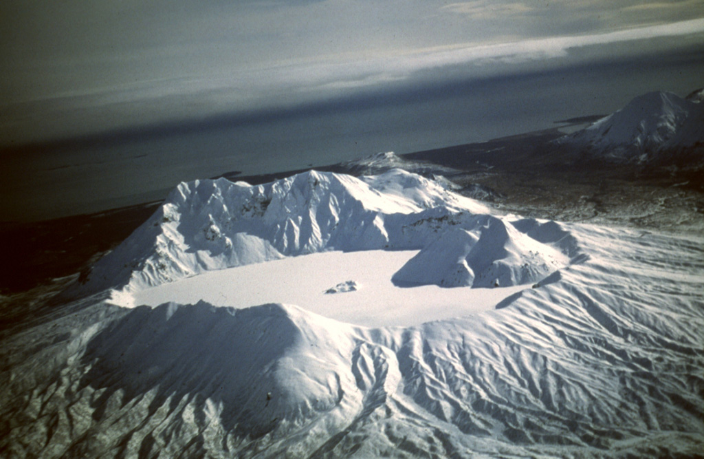 Snow and ice cover the Kaguyak caldera lake in this view from the NW. The caldera formed during a voluminous eruption about 5,800 years ago that produced pyroclastic flows that reached Shelikof Strait. Two lava domes, one large and the other seen just above the center of the lake, were built after the caldera-forming eruption. Photo by Tom Miller, 1998 (Alaska Volcano Observatory, U.S. Geological Survey).