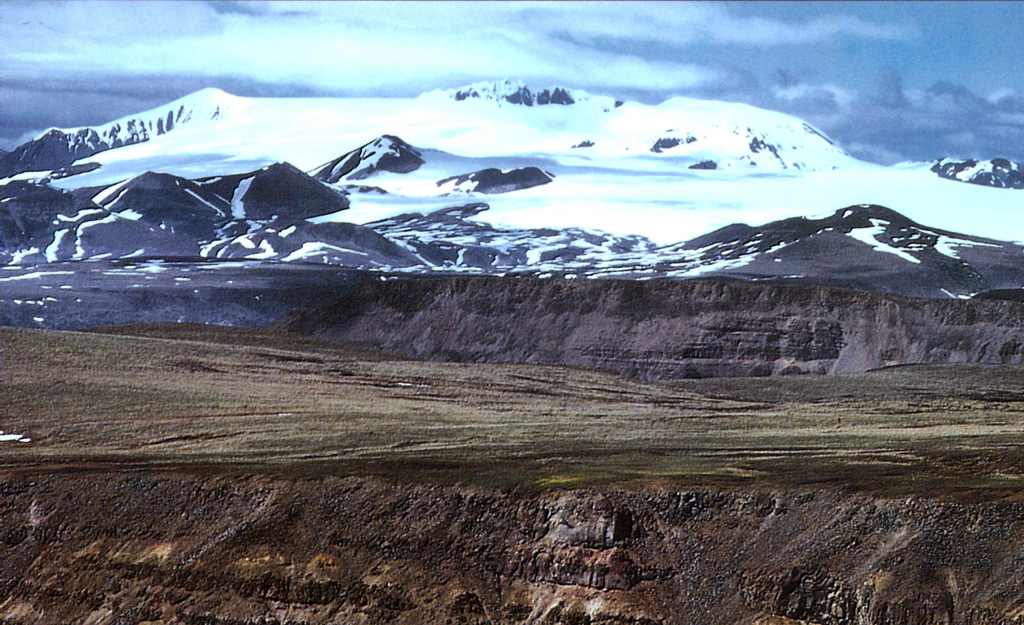 The central ice-covered summit complex of Mount Edziza is seen here from the SW rising above the Kitsu and Big Raven plateaus. Late-Tertiary lava flows are the foreground. The smaller hills at the base of the icecap are Quaternary cones. Mount Edziza contains a 2-km-wide, ice-filled caldera at its summit, and numerous ice-contact features and products of subglacial eruptions. The complex contains numerous Holocene cones, some of which are younger than about 1,300 years. Photo by Jack Souther, 1992 (Geological Survey of Canada).
