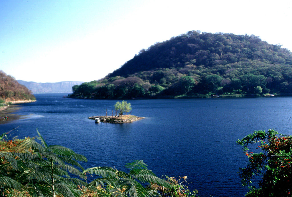 This vegetated island is Cerro Grande, a post-caldera lava dome in the SW side of Coatepeque caldera. The partially submerged dome has a basal diameter of about 2.5 km and a volume of about 0.5 km3. The smaller San Pedro Island with one tree is exposed during low lake levels. Photo by Lee Siebert, 2002 (Smithsonian Institution).