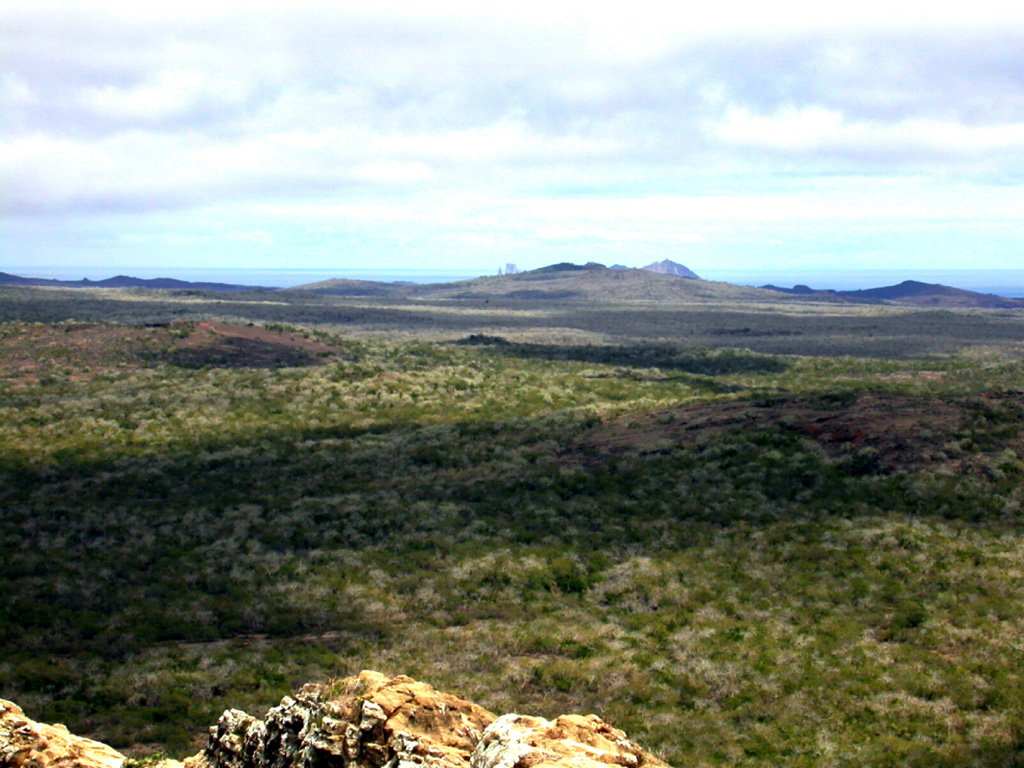 The NW coast of San Cristóbal Island contains numerous young pyroclastic cones, spatter vents, and lava flows.  The island consists of a densely vegetated SW part and a lower, younger NE part with many extremely youthful lava flows.  The tip of Kicker Rock, two steep-cliffed islands separated by a narrow cleft, is barely visible on the center horizon.  Kicker Rock is an eroded remnant of a tuff cone that forms one of the scenic highlights of the Galápagos, 5 km off the west coast of San Cristóbal. Photo by Ed Vicenzi, 2002 (Smithsonian Institution).