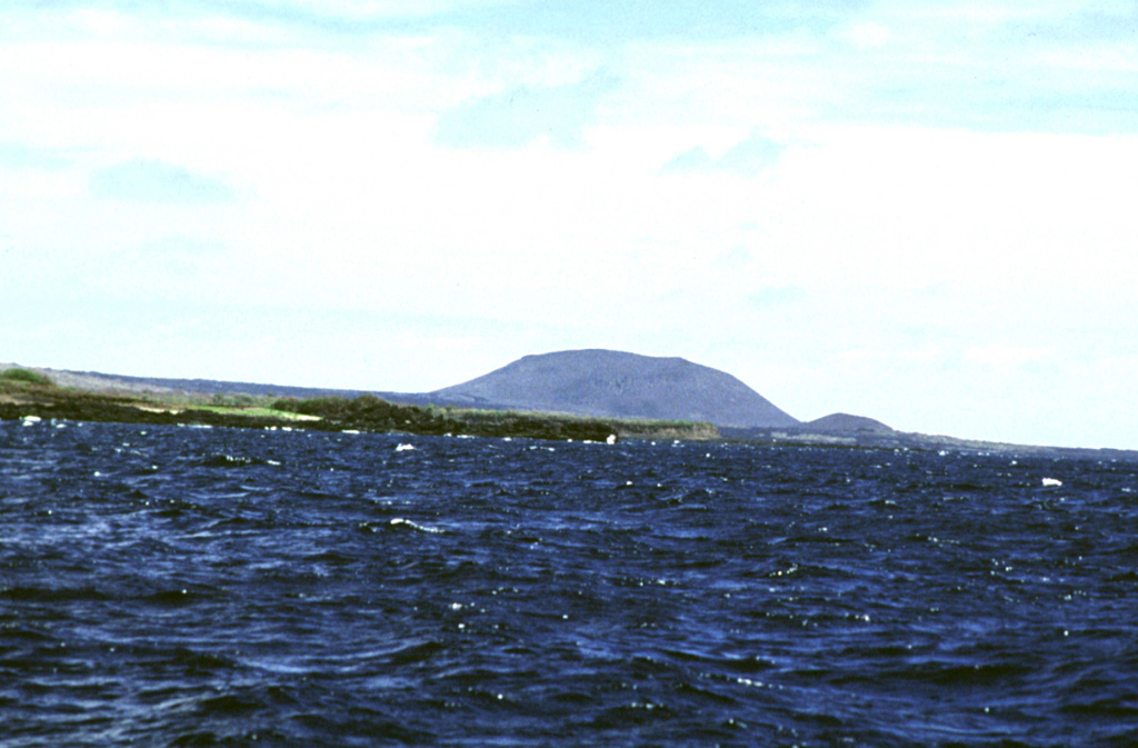 A large tuff cone rises near Punta Calle on the southern coast of Marchena Island beyond fresh-looking lava flows west of the cone in the foreground.  The low shield volcano forming Marchena Island contains one of the largest calderas of the Galápagos Islands.  In contrast to other Galápagos volcanoes, the 6 x 7 km caldera and its outer flanks have been largely buried by a cluster of pyroclastic cones and associated lava flows.  The first historical eruption of Marchena occurred in 1991.   Photo by Ed Vicenzi, 1984 (Smithsonian Institution).