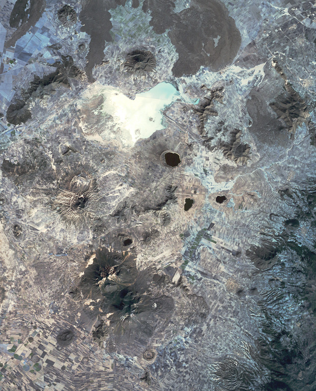 This view of the Serdán-Oriental basin covers about 38 km in a N-S (vertical) direction.  The circular peak at the upper center, partially surrounded by young lava flows from Los Humeros caldera, is Cerro Pizarro lava dome.  The lake-filled maar to the SSE across the light-colored dry lake basin is Alchichica maar.  The erosionally ribbed flanks of Cerro Xalapaxco tuff cone and Cerro Pinto lava dome lie to the SW, and the large forested peaks at the bottom are the the twin Las Derrumbadas lava domes. NASA Landsat satellite image, 1999 (courtesy of Loren Siebert, University of Akron).
