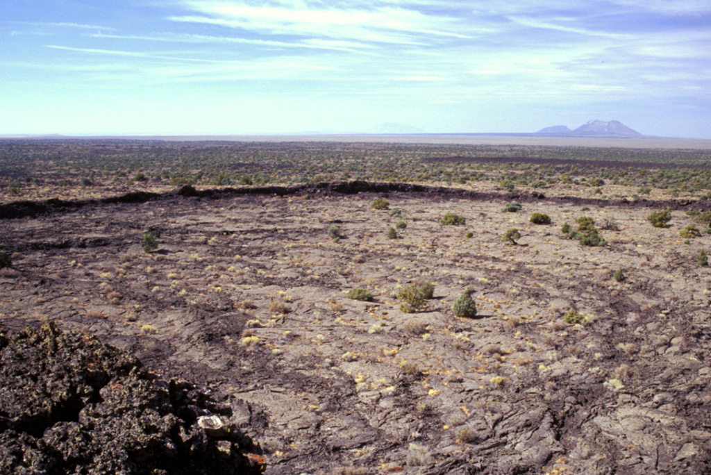 A broad frozen lava lake rises almost to the rim of the 800 x 300 m wide summit crater of Hell's Half Acre shield volcano, the easternmost of the young basaltic lava fields of the Snake River Plain.  The 300 x 800 m wide lava lake is seen from the summit of the low shield volcano, with East Butte and Middle Butte visible to the west on the upper right horizon.  Dominantly pahoehoe lava flows were erupted from a 3-km-long, NW-SE trending vent system in the NW part of Hells Half Acre lava field about 5200 years ago.  Photo by Lee Siebert, 2002 (Smithsonian Institution).