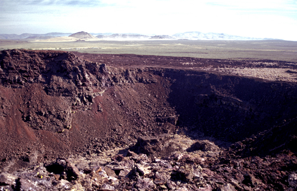 One of a series of interlocking craters forming the complex summit of Black Butte, the vent of the Shoshone lava field, is seen here from the summit of the butte.  The broad, low Shoshone shield volcano fed voluminous lava flows that traveled a small distance north towards the Mount Bennett Hills in the background, but the bulk of the flows traveled initially south and then west for a total distance of 60 km.  The Shoshone lava field, erupted about 10,000 years ago, is the westernmost of the young volcanic fields of the Snake River Plain. Photo by Lee Siebert, 2002 (Smithsonian Institution).