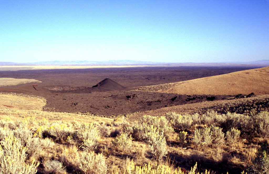 Coffeepot Crater (left of the cone) was the source of voluminous basaltic lava flows in the Jordan Craters volcanic field of SE Oregon. The roughly 200-m-wide crater lies at the NW end of the 1.6 km3 lava field. A WSW-ENE-trending line of spatter cones in the right foreground extends towards the crater. The lava field forms one of Oregon's youngest lava flows at less than 3,200 years old. Photo by Lee Siebert, 2002 (Smithsonian Institution).