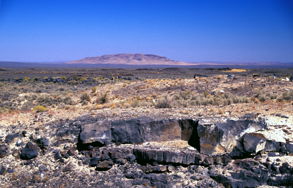 Basaltic lava flows from Saddle Butte volcanic field in SE Oregon descend low-angle slopes towards Saddle Butte on the horizon.  The volcanic field consists of an 1100 km2 area of Pleistocene basalts with a superposed 240 km2 area of younger Pleistocene basalts.  Oregon State Highway 78, from where this photo was taken, cuts across the western side of the isolated lava field, which reaches nearly to the southern base of Saddle Butte.  Lava tubes are prominent in the younger flows. Photo by Lee Siebert, 2002 (Smithsonian Institution).