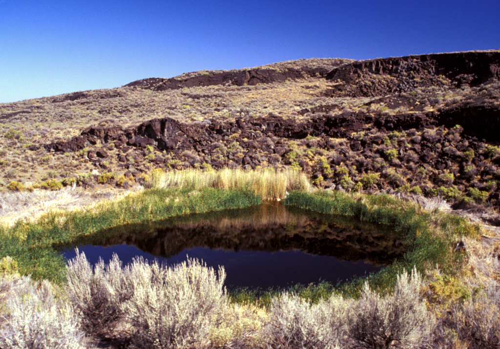Lava flows of West Dome, one the structural highpoints of Diamond Craters, rise NE of lake-filled Malheur Maar. The shallow 2-m-deep lake occupies one of many maars (the rest of which are dry) of the Diamond Craters volcanic field at the time of this 2002 photo. Diamond Craters consists of a 70 km2 area of basaltic lava flows, scoria cones, and maars. The initial eruptions of pahoehoe lava flows and later activity involved magma injection that produced six structural highs of up to 150 m. Photo by Lee Siebert, 2002 (Smithsonian Institution).
