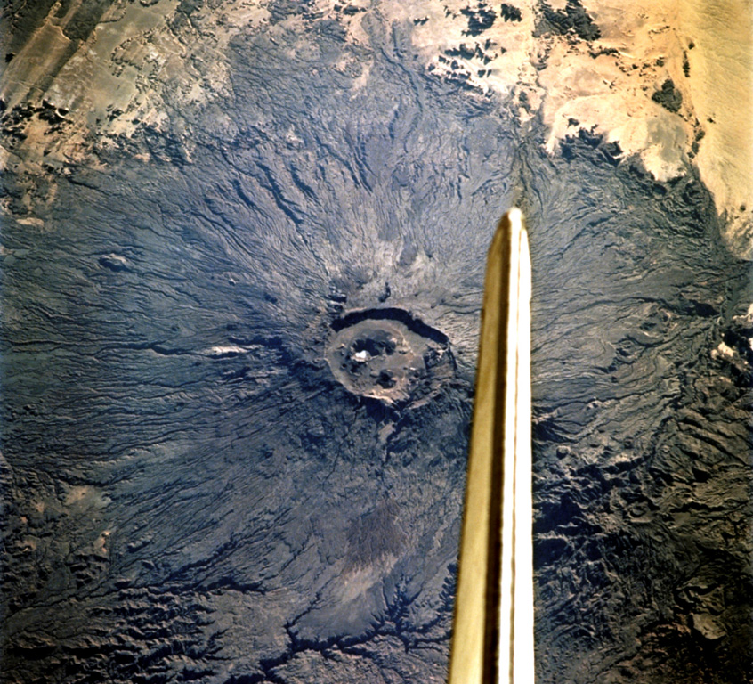 The vertical stabilizer of the Space Shuttle Endeavor appears above the summit caldera of Emi Koussi volcano at the SE end of the Tibesti Range in Chad. Nested 12 x 15 km wide calderas at the summit contain large craters and young basaltic scoria cones. NASA Space Shuttle image STS108-701-008, 2001 (http://eol.jsc.nasa.gov/).