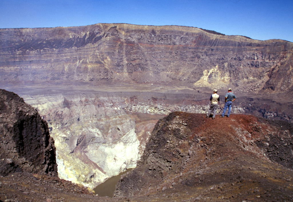 Geologists on the southern rim of Santa Ana's summit crater stand above the hydrothermally altered walls of the more than 100-m-deep inner crater. A near-vertical fault is visible in the far northern wall of the larger crater beyond the crater floor in the center of the photo, above the inner crater. Lava flows exposed in the crater wall are overlain by a roughly 10-m-thick light-brown sequence of phreatomagmatic tephra layers. Photo by Paul Kimberly, 2002 (Smithsonian Institution).