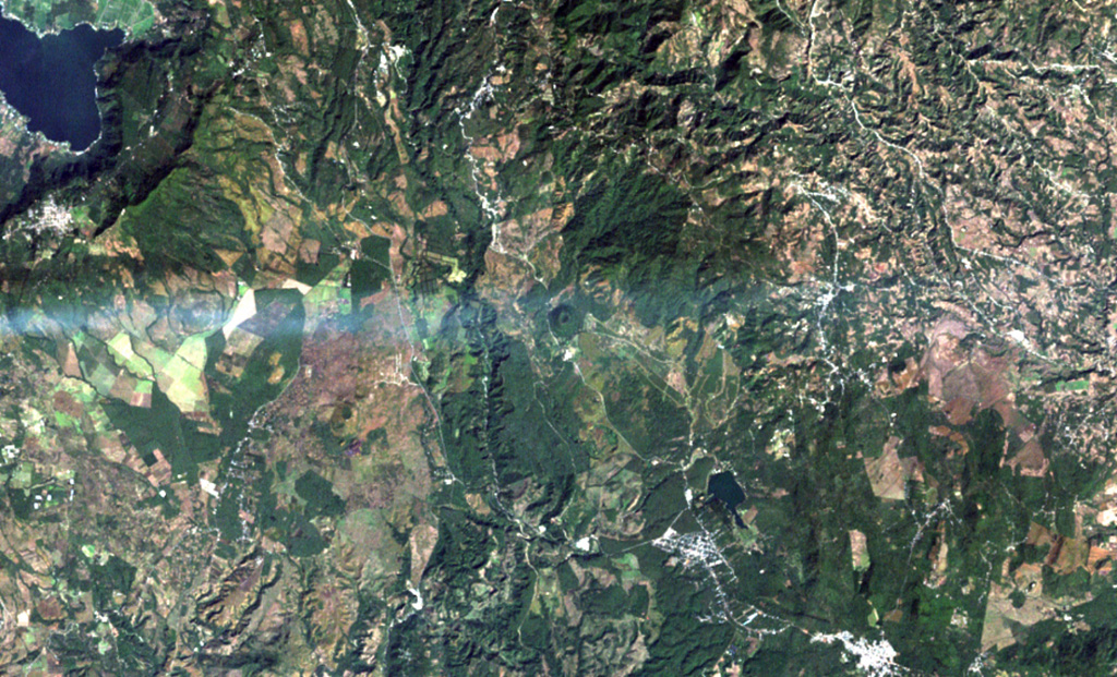 A volcanic plume from Pacaya volcano extends eastward from the left to Cerro Redondo, a small symmetrical cinder cone in the center of the image.  This cone is one of the youngest features of the Cuilapa-Barbarena volcanic field, a cluster of about 70 Quaternary cinder cones located near the cities of Cuilapa and Barbarena (lower right) and in areas to the north and west.  The cones were constructed where the NW-SE-trending Jalpatagua Fault intersects the southern margin of the Miocene Santa Rosa de Lima caldera. NASA Landsat image, 2000 (courtesy of Loren Siebert, University of Akron).