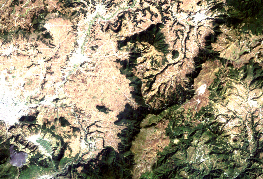 The triangular dark-green area (top-center) is the Cuxliquel lava-dome complex, lying within a semi-arcuate depression near the city of Totonicapán (upper right).  The small green circle below and to the right of the larger dome complex is Cerro de Oro, a small symmetrical lava dome.  The Río Samalá traces an arcuate path around the Quaternary dome complex beginning on its SE side (center) and progressing around to the north and then to the SW (bottom left).  The white-colored area (left-center margin) is the city of Quetzaltenango. NASA Landsat image, 2000 (courtesy of Loren Siebert, University of Akron).