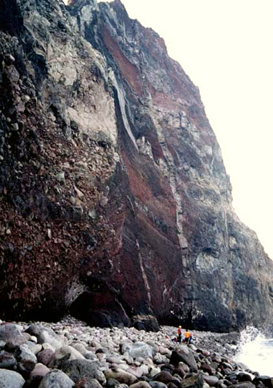 Geologists stand on the narrow cobble beach at the NE base of Sumisujima. Vertical light-colored dikes cut intrusive rocks at the lower right alogn with oxidized tephra layers, breccias, and lava flows forming the pinnacle. The spire is an eroded remnant of the pre-caldera volcano on the outer southern flank of a large submarine caldera. A large segment of the eastern side of the pinnacle previously slumped into the sea. Copyrighted photo by Kenichiro Tani, 2002 (Japanese Quaternary Volcanoes database, RIODB, http://riodb02.ibase.aist.go.jp/strata/VOL_JP/EN/index.htm and Geol Surv Japan, AIST, http://www.gsj.jp/).