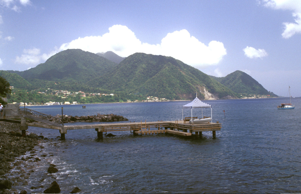 The Plat Pays volcanic complex rises to the south beyond the shoreline of the city of Roseau in the foreground.  Three lava domes that formed after the roughly 39,000-year-old caldera collapse at Plat Pays lie on the horizon.  On the left is the rounded Morne Canot dome, in the center is La Falaise dome, and the peak at the far right is Morne la Vue, which rises above the village of Pointe Michel.  The summit of the Morne Plat Pays stratovolcano lies in the left-center background between Morne Canot and La Falaise domes. Photo by Lee Siebert, 2002 (Smithsonian Institution).