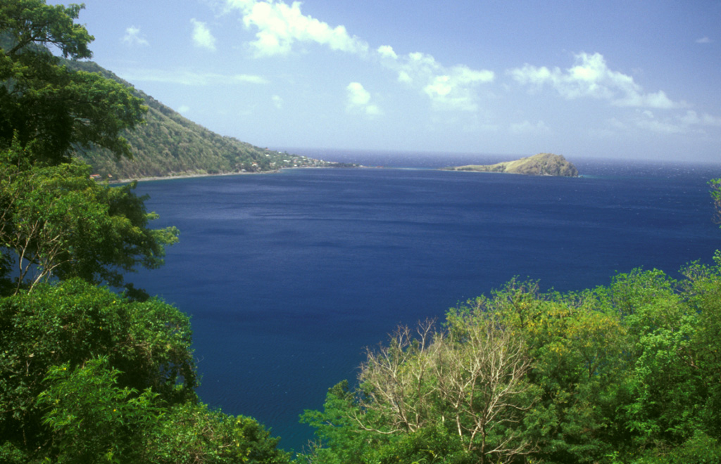 Scotts Head lava dome forms a dramatic promontory marking the southern margin of Soufrière Bay and is connected by a narrow spit only a few meters high to Scotts Head village (left-center).  The weathered hill (also known as Pointe Cachacrou) consists of subvertical lava sheets and is a remnant of lava dome extruded along a fracture inboard of the southern margin of the Soufrière Bay depression.  The slopes of Crabier lava dome rise at the left. Photo by Lee Siebert, 2002 (Smithsonian Institution).