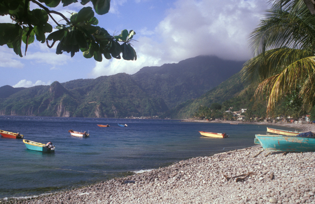 The summit of the steep-sided Sorcière lava dome rises from the coast at the left to just above the horizon.  Morne Acouma lava dome (left-center) lies to the right of Sorcière in this view from Soufrière harbor (south of the domes) and was the source of a lava flow that extends to the northern edge of the town.  Coastal cliffs below the domes consist of block and ash flow deposits associated with growth of the domes.  The dacitic Morne Acouma dome is older than about 28,500 years. Photo by Lee Siebert, 2002 (Smithsonian Institution).