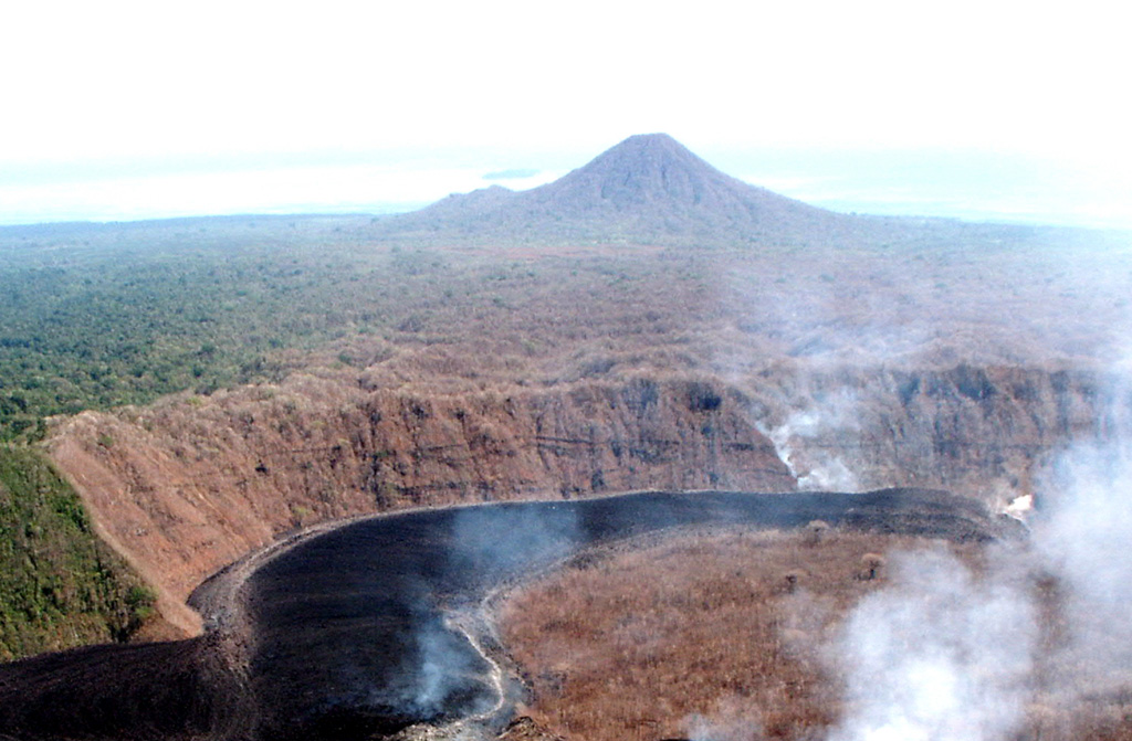 Lolo rises to the N beyond lava flows producing gas plumes during the 2002 eruption of Pago. Lolo is located N of Pago volcano along the N coast of New Britain, near Cape Hoskins. It is of late Pleistocene or Holocene age and overlaps the older Kapberg volcano on its W side. Lola contains a 250-m-wide and 60-m-deep crater.  Photo by Elliot Endo, 2002 (U.S. Geological Survey).