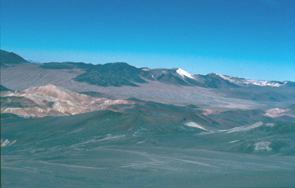 Cerro Escorial (center horizon), viewed from the summit of Lastarria volcano, is a small andesitic-dacitic stratovolcano that straddles the Chile/Argentina border.  Very youthful-looking lava flows of uncertain age are seen here extending 3-4 km SW-ward over an ignimbrite deposit on the Chilean side of the border.  Cerro Escorial is located 4 km NE of an active sulfur mine in older, extensively hydrothermally altered rocks, some of which are seen in the middle ground.    Photo by José Naranjo, 1983 (Servico Nacional de Geologica y Mineria).