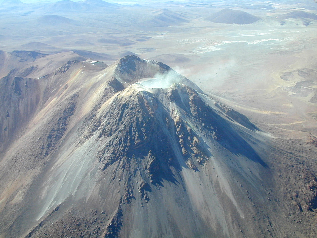 Steam rises from the fumarolically active southern summit crater of Irruputuncu, a small stratovolcano that straddles the Chile/Bolivia border.  Irruputuncu, seen here from the WSW, was constructed within the collapse scarp of a Holocene debris avalanche whose deposit extends to the SW.  Levees of viscous lava flows down the western flank of an edifice that was constructed within this scarp are seen at the lower left.  The first unambiguous historical eruption from Irruputuncu took place in November 1995. Photo by José Naranjo, 2001 (Servico Nacional de Geologica y Mineria).