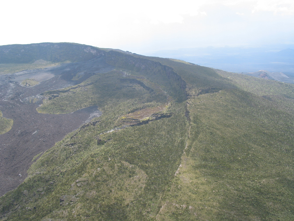 A fissure (center) cuts the SE caldera rim of Nyamuragira. Several historical eruptions have occurred from both summit and flank vents and have produced lava flows that covered portions of the caldera floor and traveled long distances down the flanks of the volcano. Photo by Simon Carn, 2004 (TOMS Volcanic Emissions Group, University of Maryland, Baltimore County).