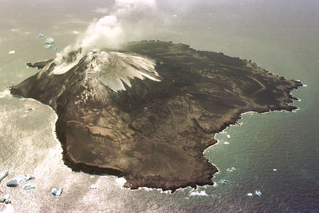 Steam clouds rise from fumaroles near the summit of Mount Curry, on Zavodovski Island, in this aerial view from the south. Icebergs lie off the shores of the approximately 4.5-km-wide island, the northernmost of the South Sandwich Islands. The eastern coast (right) comprises lava flows and is low-lying, contrasting with the cliffs of the west (left). Two young eruptive fissures extend NE from the summit toward craters on the east flank and were the source of a lava platform extending toward Pungent Point (right). Photo by HMS Endurance (courtesy of John Smellie, British Antarctic Survey).