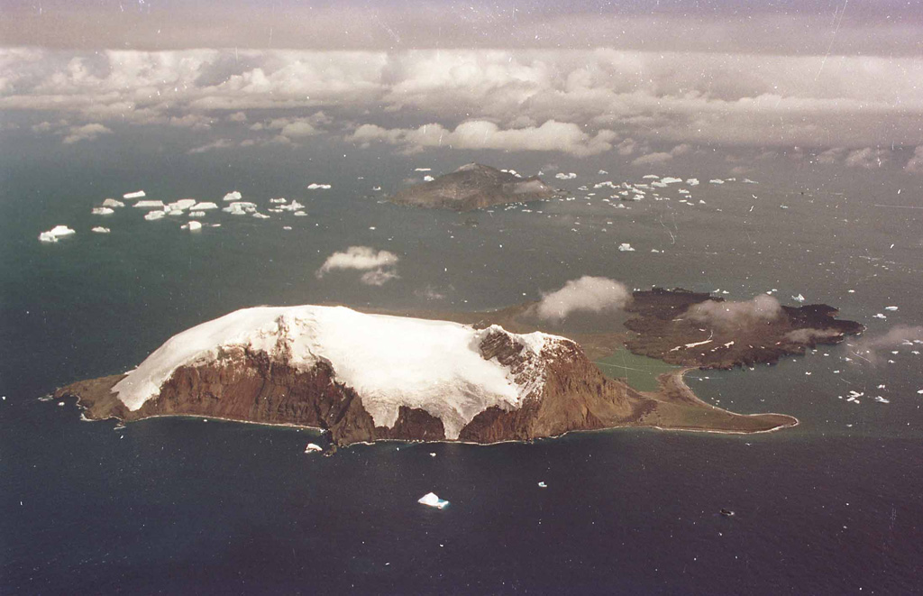 The glacier-covered peaks of Mount Andromeda (Ieft) and Mount Perseus (right) rise along the eastern side of Candlemas Island. Rocky beaches join this prominent southern part of the island to a low-relief ice-free area consisting of dark lava flows from Lucifer Hills in the north. Lucifer Hills is a complex of coalesced cinder cones with numerous active fumaroles, and dark clouds were seen emanating from the area in 1823 and 1911, indicating recent eruptions. A narrow spit, Demon Point, divides greenish Gorgon Pool from Kraken Cove (right). Medusa Pool lies below the cloud beyond Gorgon Pool. Vindication Island can be seen in the background, about 4.5 km from Candlemas. These two islands are part of the same volcanic edifice, with Vindication Island representing the oldest parts of the volcano. Photo by HMS Endurance (courtesy of John Smellie, British Antarctic Survey).