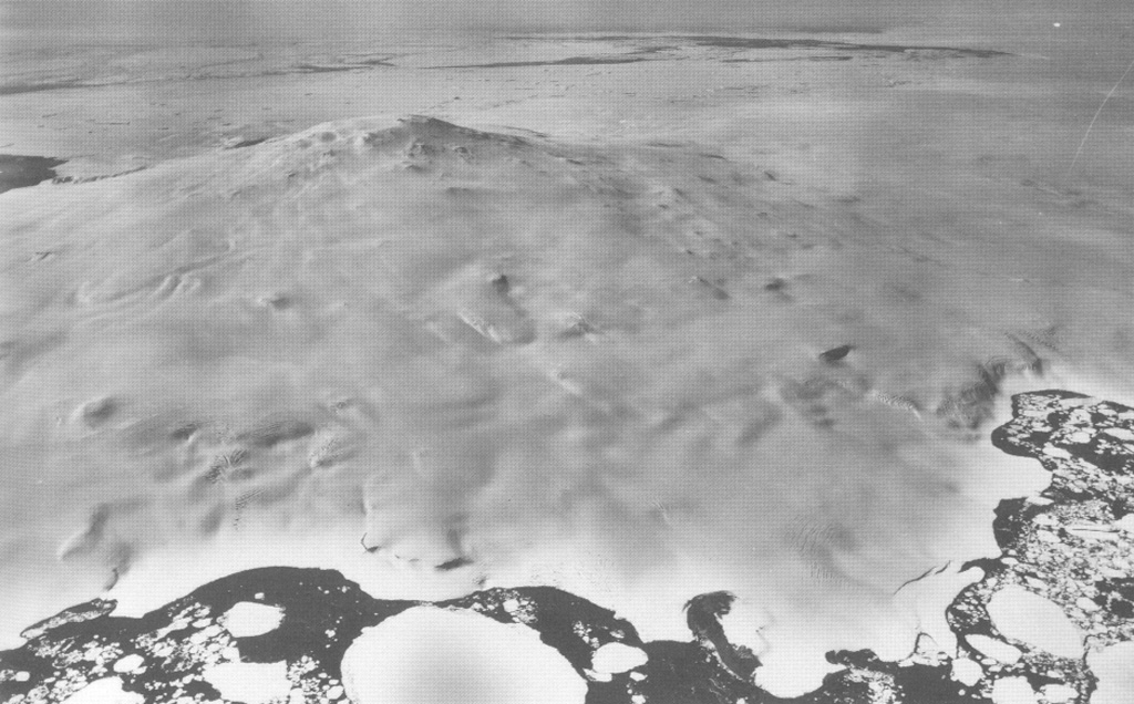 Mount Siple forms the high point of Siple Island in Marie Byrd Land off the coast of Antarctica. This aerial view looks east, with dark-colored open water in the foreground and the Getz Ice Shelf between the island and Antarctica. Most volcanic features are buried in snow and ice, but the Lovill Bluff tuff cone outcrops at sea level (bottom right). Trachytic rocks at the summit have been Ar-Ar dated to about 227,000 and 169,000 years old. U. S. Navy photo TMA 1627 F33 088, 1985.