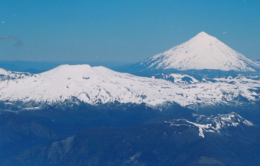 Quetrupillan stratovolcano (left) lies at the center of a group of three volcanoes trending transverse to the Andean chain.  It is seen here from the summit of Villarrica volcano (at the western end of the chain), with conical Lanín volcano at the eastern end in the background.  The 2360-m-high Quetrupillan volcano was constructed within a large 7 x 10 km wide caldera; a smaller caldera truncates the summit.  Some of the most recent activity produced pyroclastic cones along the right-hand flank, near the SW margin of the older caldera.    Photo by Judy Harden, 2004 (University of South Florida).