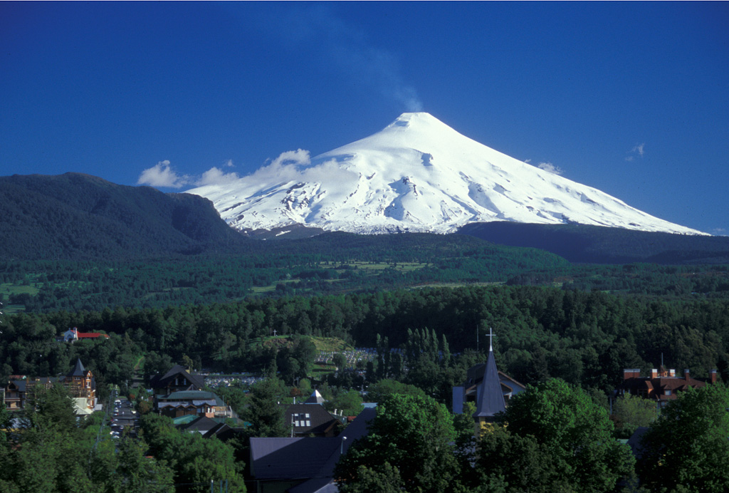 Snow-covered Villarrica, one of Chile's most active volcanoes, rises above the resort town of Pucón below its northern flank.  A faint steam plume drifts from an active lava lake in the summit crater.  The steep summit cone was constructed within a mostly buried, 2-km-wide caldera whose dissected outer flanks rise above the tree line.  Villarrica is the westernmost of three large stratovolcanoes that trend perpendicular to the Andean chain.  Historical eruptions have been documented since 1558 CE. Photo by Lee Siebert (Smithsonian Institution). 2004.