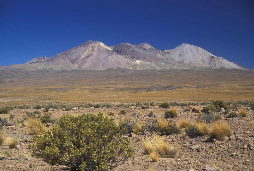 The elongated Taapaca massif rises to the SE above the gentle slopes of block-and-ash flow deposits from the volcano.  The steeply dipping lava flow on the left horizon caps hydrothermally altered rocks of a Pleistocene stratovolcano of the Taapaca II complex.  The dome complex at the center is of part of the dacitic Pleistocene Taapaca III complex, and the light-colored dome at the right is part of the dacitic Pleistocene-to-Holocene Taapaca IV complex. Photo by Lee Siebert, 2004 (Smithsonian Institution).
