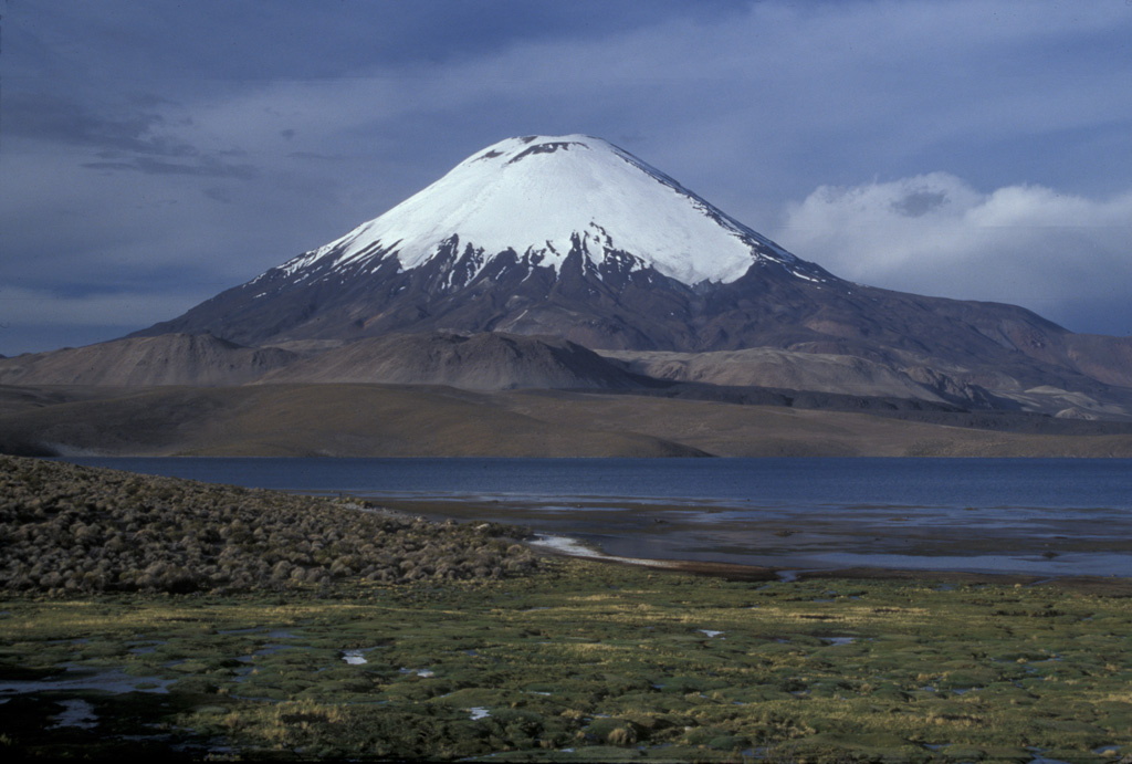 Glacier-clad Volcán Parinacota rises to the NE above Laguna Chungará near the Chile-Bolivia border.  The lake was formed when collapse of Parinacota about 8000 years ago produced a 6 cu km debris avalanche that traveled 22 km to the west and blocked drainages.  Subsequent eruptions constructed the 6348-m-high symmetrical stratovolcano, which towers above late-Pleistocene andesitic-to-rhyolitic lava domes and flows in the middle ground.    Photo by Lee Siebert, 2004 (Smithsonian Institution).
