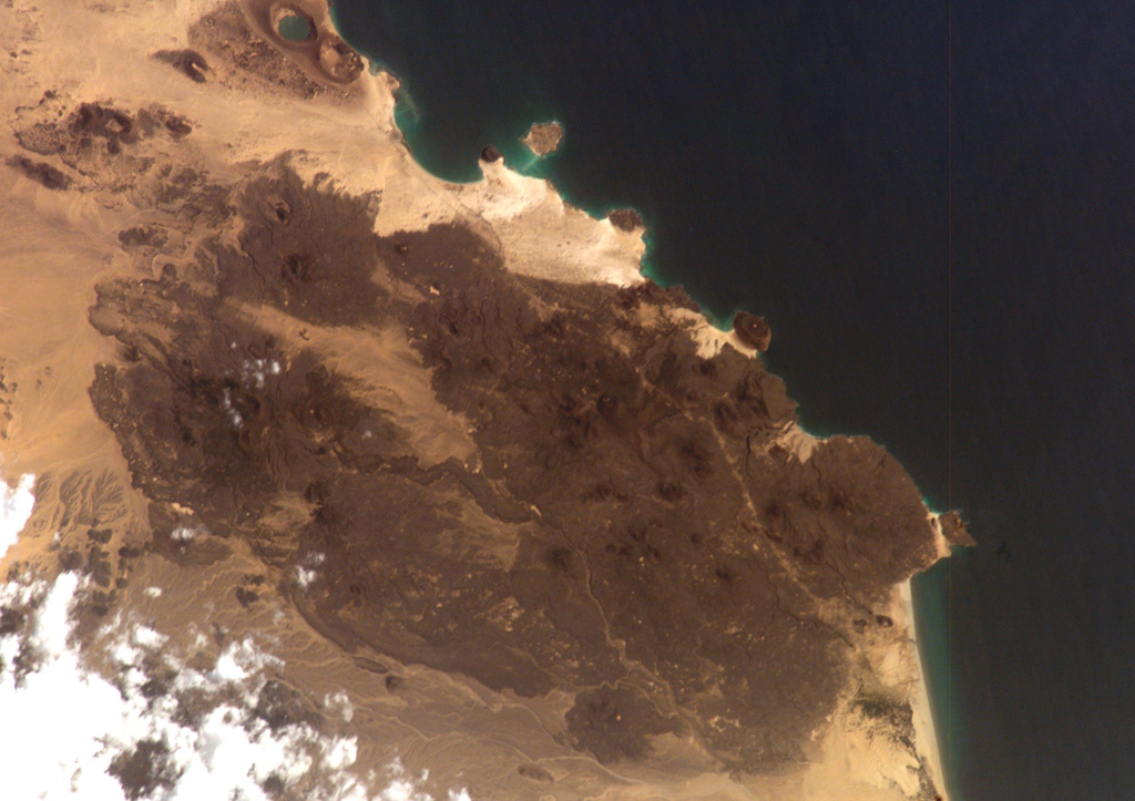 The Harra of Bal Haf volcanic field fills much of this NASA International Space Station image taken along the Gulf of Aden in southern Yemen (N to the bottom left). Dark-colored lava fields cover much of the volcanic field and create an irregular shoreline. One basaltic flow may be of historical age. Two large tuff cones are visible along the coast at the upper left, one of which is lake filled. NASA International Space Station image ISS006-E-5153, 2002 (http://eol.jsc.nasa.gov/).