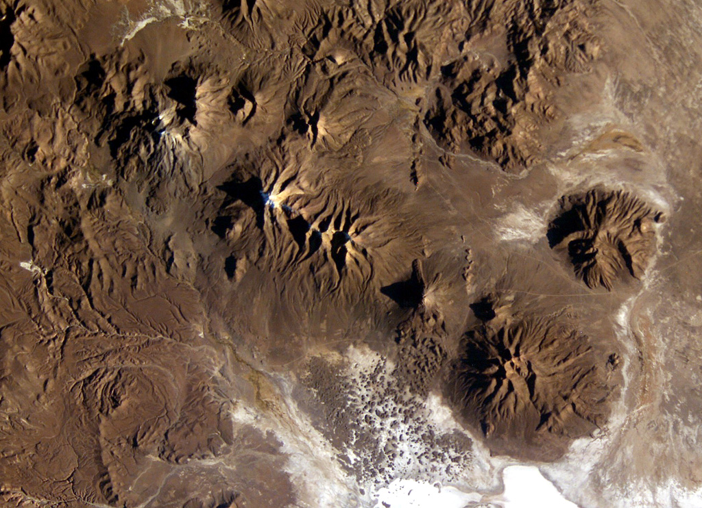 A long E-W-trending volcanic chain extends across the border between Chile and Bolivia in this NASA International Space Station image (with north to the upper right).  The chain extends from historically active Isluga volcano (upper left) to eroded Saxani volcano at the lower right. The smaller volcano immediately to the west of Saxani with a sharp shadow is the steep-sided Tata Sabaya volcano.  Tata Sabaya was the source of a major debris-avalanche deposit (bottom center) that forms the small dark-colored hills on the white floor of Salar de Coipasa. NASA International Space Station image ISS009-E-6849, 2004 (http://eol.jsc.nasa.gov/).