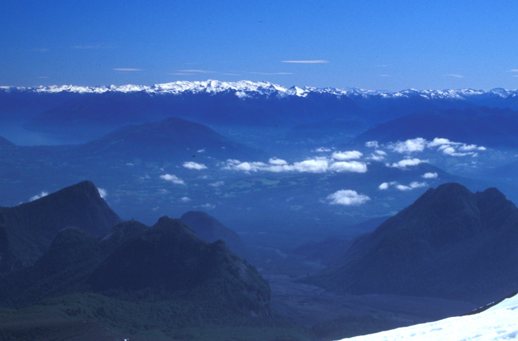 The broad profile of the snow-covered Sollipulli massif lies on the horizon to the NE as seen from the upper slopes of Villarrica volcano.  A 4-km-wide caldera with post-caldera lava domes on its rim lies on the eastern side of the Nevados de Sollipulli volcanic chain.  The rounded hills in the middle distance are pyroclastic cones of the Caburgua-Huelemolle volcano group; lava flows from these cones dammed drainages, forming Laguna Caburgua, visible at the upper left. Photo by Lee Siebert, 2004 (Smithsonian Institution).
