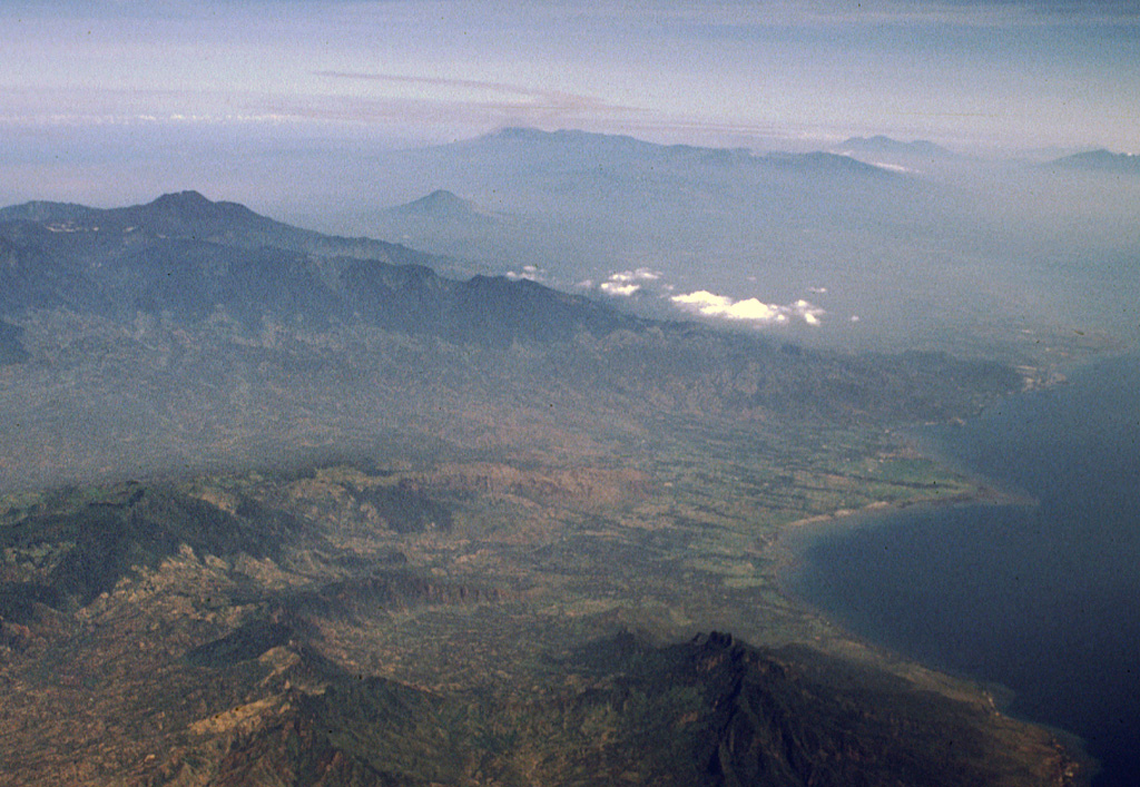 The small Lurus volcanic complex (middle right) lies along the northern coast of eastern Java, below and to the right of the low clouds at the right-center. Lurus lies N of the Iyang-Argapura massif, the broad volcanic complex covering much of the left center of this aerial view from the NE. The conical volcano beyond Iyang-Argapura is Lamongan volcano, and the Tengger-Semeru massif lies along the center of the horizon. The triple-peaked volcano on the right horizon is Kawi-Butak. Photo by Lee Siebert, 2000 (Smithsonian Institution)