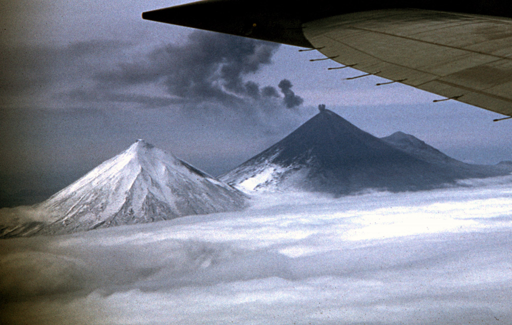 A small ash plume rises from the summit crater of Pavlof volcano on 28 May 1960 as the detached plume from an earlier explosion drifts away to the east. Ash deposits are visible on the flanks in this view from the north. Mild ash eruptions took place from about 1960 to 1963, especially during July 1962 to June 1963. The peak to the left is Pavlof Sister, and Little Pavlof forms the smaller peak to the right of Pavlof. Photo by Ken Morin, 1960 (courtesy of Bill Rose, Michigan Technological University).