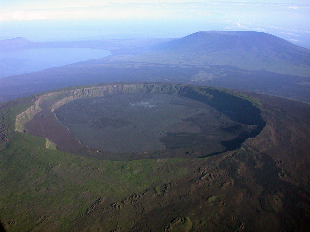 A spectacular aerial view from the SE shows the caldera of Darwin volcano in the foreground, with Volcán Wolf in the right background and the tip of Volcán Ecuador at the NW tip of Isabella Island on far left horizon.  Volcán Darwin, named after the renowned naturalist, contains a symmetrical 5-km-wide, 200-m-deep summit caldera whose floor is nearly covered by youthful lava flows.  A broad terrace occupies the SW part of the caldera (left side).  Fresh-looking, dark-colored lava flows from flank fissures are visible between Darwin and Wolf volcanoes.  Photo by Patricio Ramon, 2003 (Instituto Geofisca, Escuela Politecnica Nacional).
