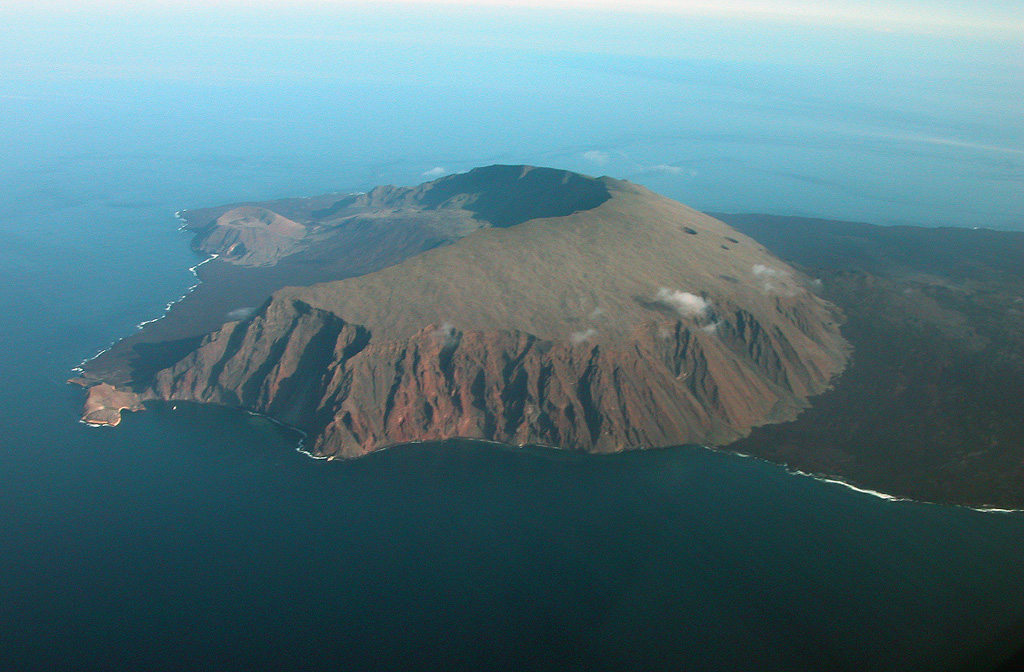 An aerial view of Volcán Ecuador from the south shows the large horseshoe-shaped caldera breached to the west that was formed when the volcano collapsed, producing a large submarine debris avalanche.  The volcano straddles the equator at the NW end of Isabela Island.  Two large pyroclastic cones were constructed along the coast, and smaller cones are found on the caldera floor.  Extensive dark-colored lava flows (right) originate from a NE-trending line of fissures that extends from the outer eastern flanks of the main edifice.  Photo by Patricio Ramon, 2005 (Instituto Geofisca, Escuela Politecnica Nacional).