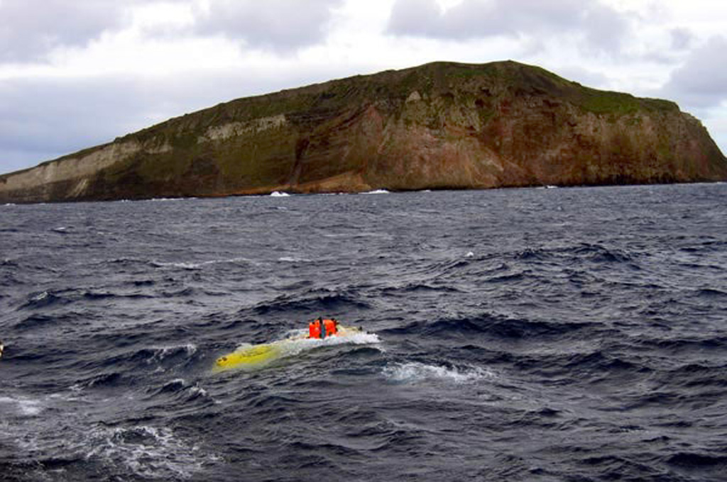 The submersible vessel Pisces V surfaces in the foreground after a dive in front of Macauley Island during a 2005 New Zealand-American NOAA Ocean Explorer research expedition to the Kermadec Arc. This view from the NW shows a prominent white band in the cliff face that is made of dacite pyroclastic flow deposits. The 3-km-wide Macauley Island is a remnant of the rim of a large submarine caldera centered 8 km to the NW and has a low, gently sloping surface truncated by steep cliffs. Image courtesy of NOAA, 2005 (http://oceanexplorer.noaa.gov/explorations/05fire).